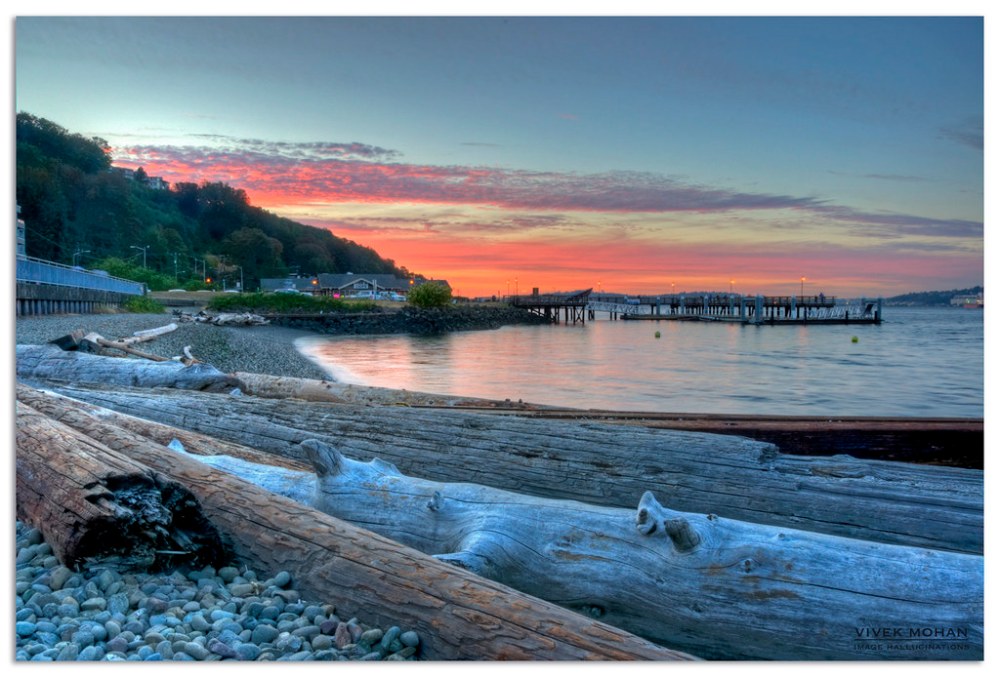 Beach at sunset with logs in foreground and pier in background under colorful sky.