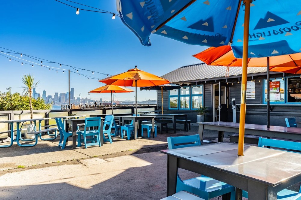 Outdoor dining area with orange umbrellas, blue chairs, city skyline in the background under a clear blue sky.