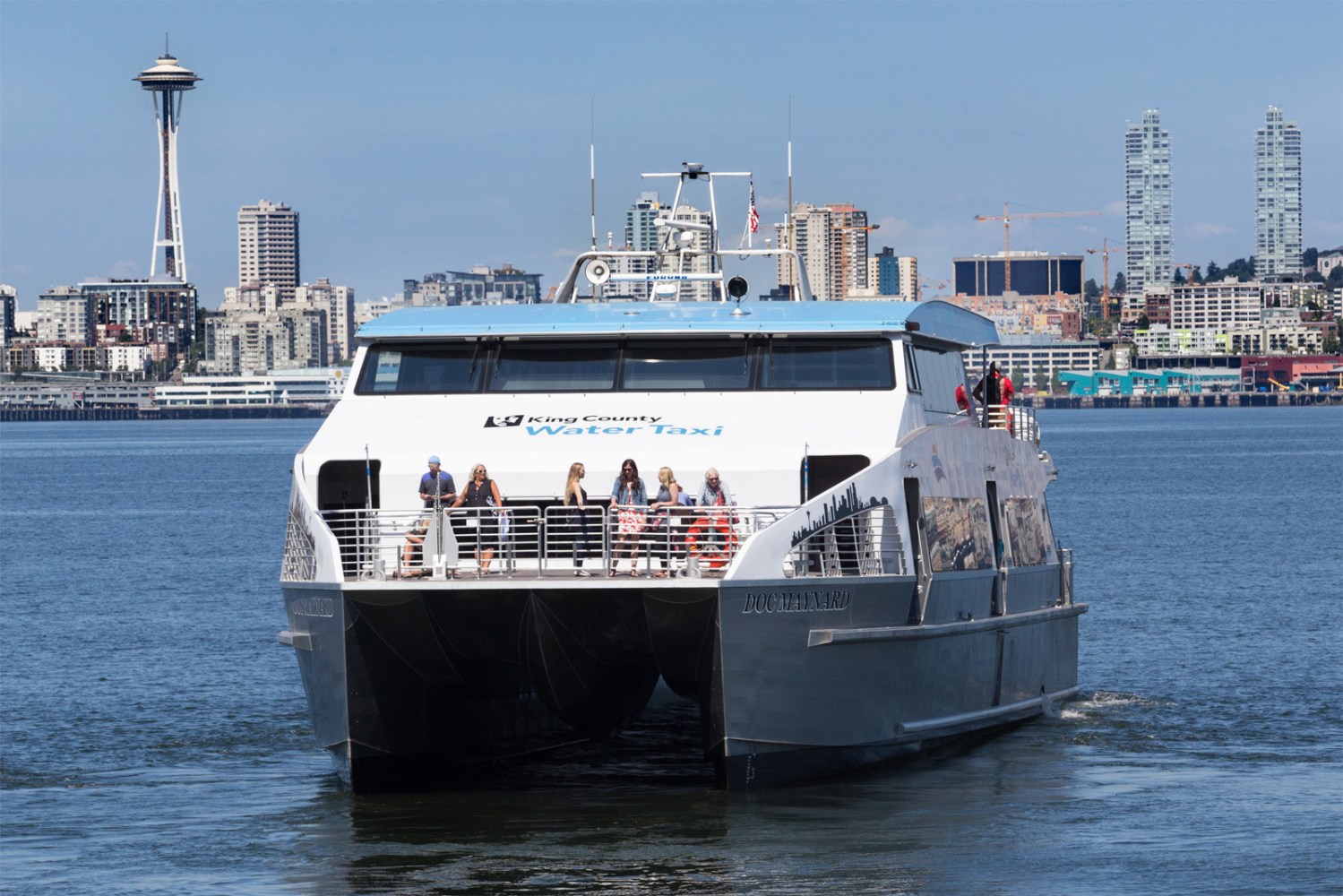 A water taxi on a city waterfront with a skyline and tower in the background.