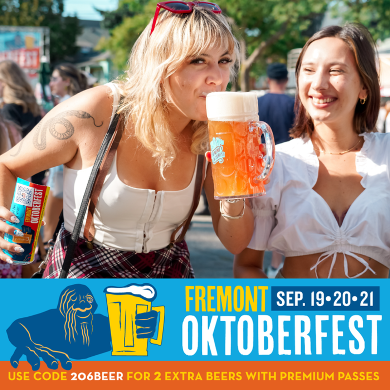 Two women enjoy large beers outdoors at Fremont Oktoberfest, with a banner advertising the event.