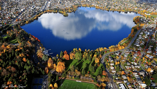 Aerial view of a lake with cloud reflection, surrounded by autumn trees and houses.